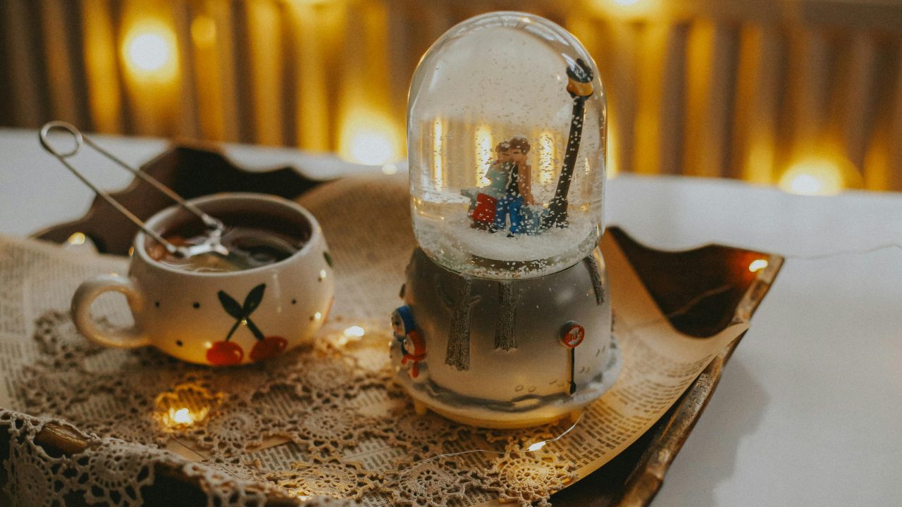 A warm winter scene featuring a snow globe and a cup of hot cocoa on a lace-covered tray with soft holiday lights in the background.